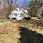 This is a before photo prior to a spring clean up service in Goffstown NH. This shows leaves that have been mulching under the snow scattered over the front lawn of a home. A wishing well is on the right side of the yard and a walkway leads up to the front porch.