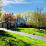 Recently completed landscaping in Hooksett NH. Large lawn and driveway are set on a hill with a blue house at the top. There are three trees in front of the home with freshly laid mulch.