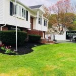 Landscaping job recently performed in Manchester NH, this photo is taken from the left side of the house where your'e able to see the beautiful shrub trimming, flowers in the front yard, dark mulch and freshly mowed lawn. They also have flowers on their front steps and a flag pole.