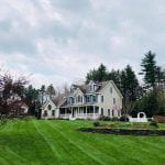 Recently completed bedford nh landscaping service. This is a wide shot of a home in bedford. Grass is bright and trimmed. There is a raised flower bed in the yard and one tree.